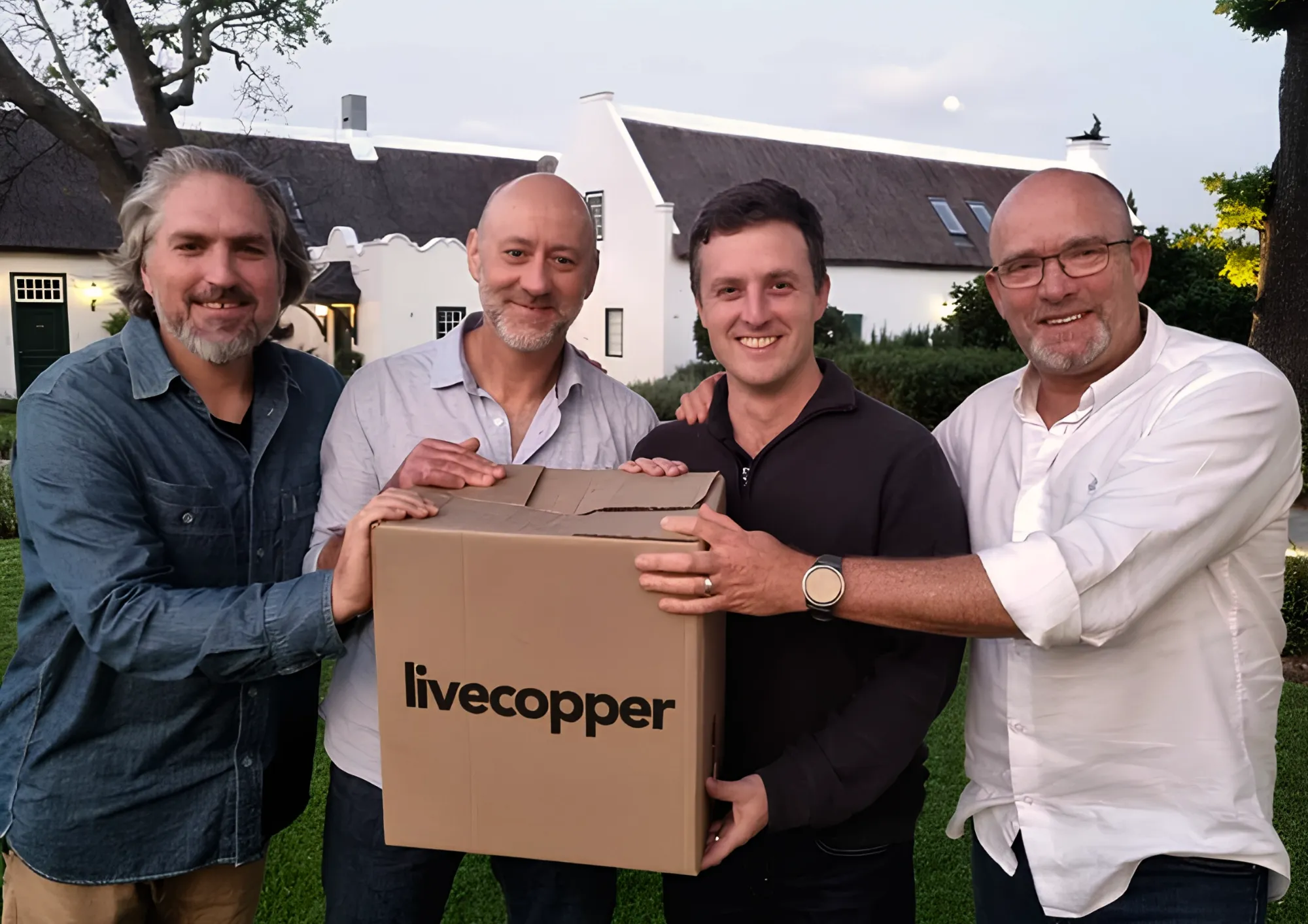 Four men holding a cardboard box printed with the Livecopper logo, in front of a thatched, whitewashed house