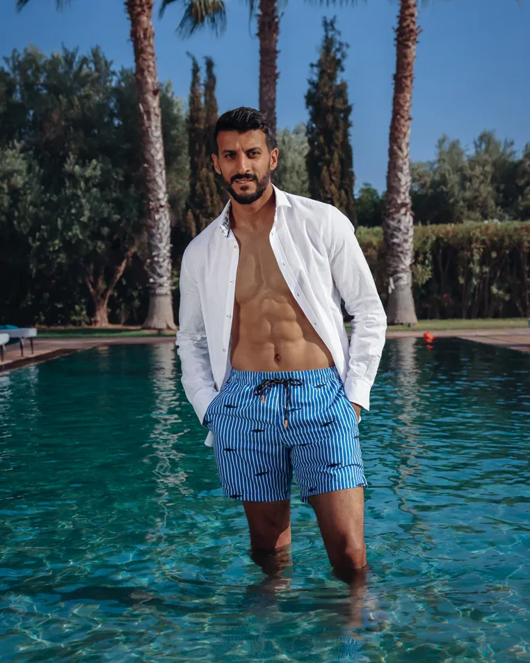 A man in A Fish Named Fred swim shorts and a white linen shirt, standing in a pool
